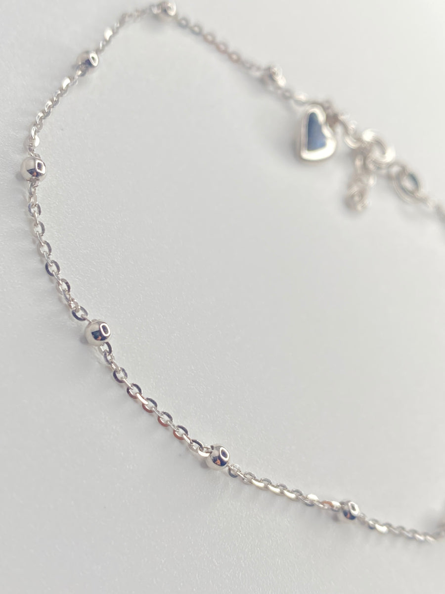 A delicate sterling silver chain anklet with small bead accents and a small heart charm, displayed on a light gray background.