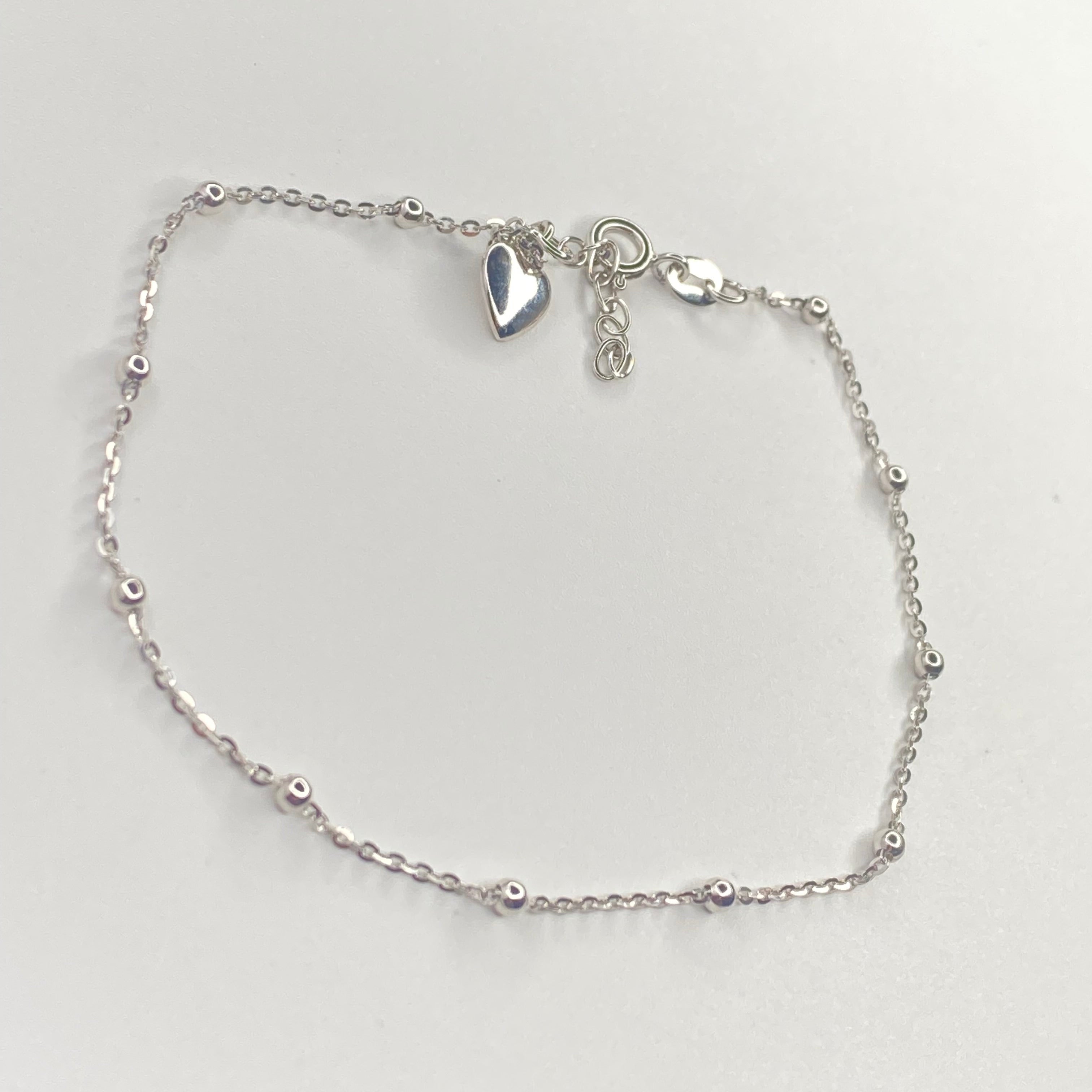 A delicate silver chain anklet with small bead accents and a small heart charm, displayed on a light gray background.