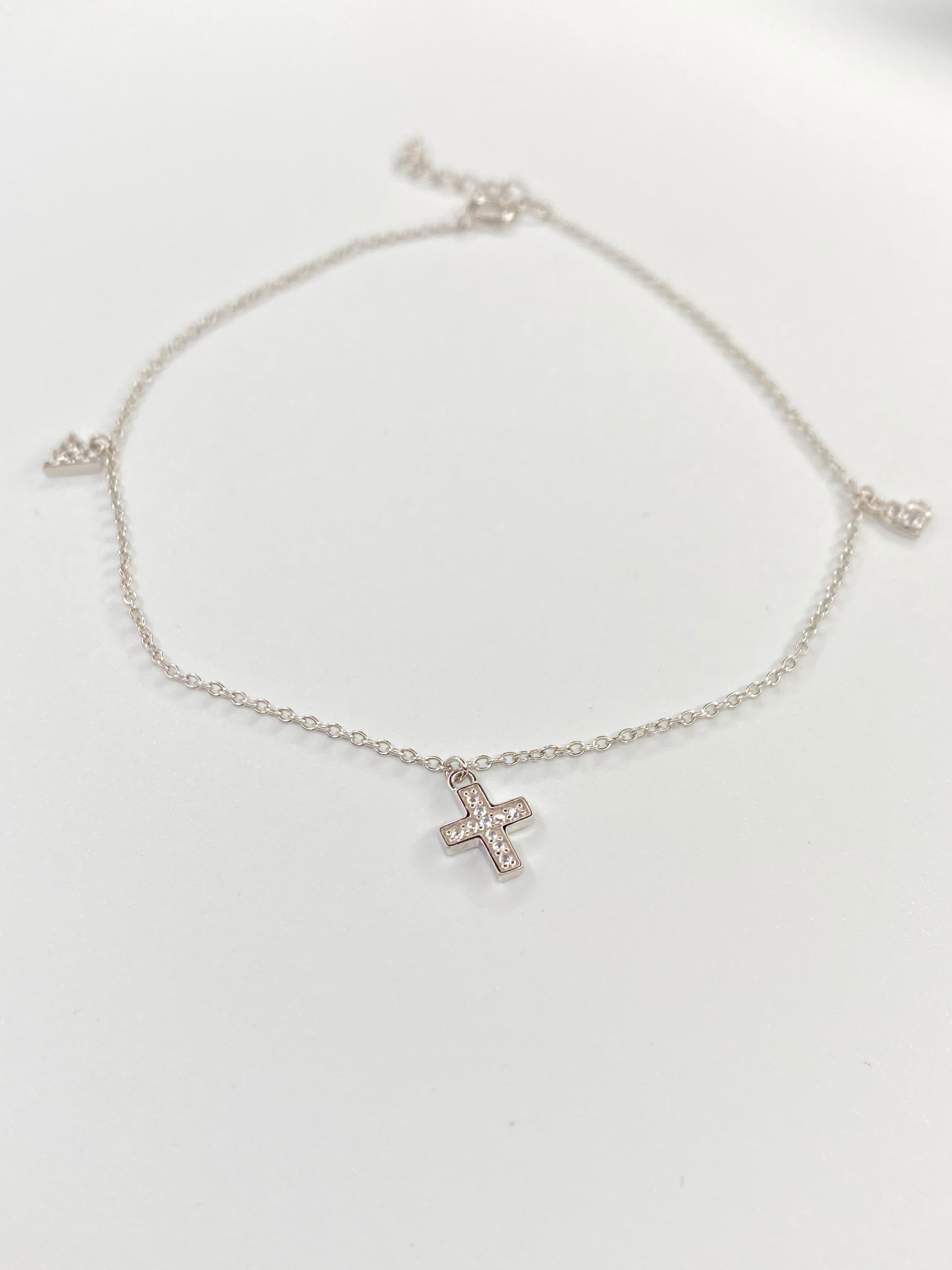 A delicate silver chain ankelt featuring a small cross charm set with clear stones, displayed on a clean white background.