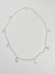 A delicate silver chain anklet with small dangling charms, including a round bezel-set iridescent stone and several clear stones, displayed on a light gray background.
