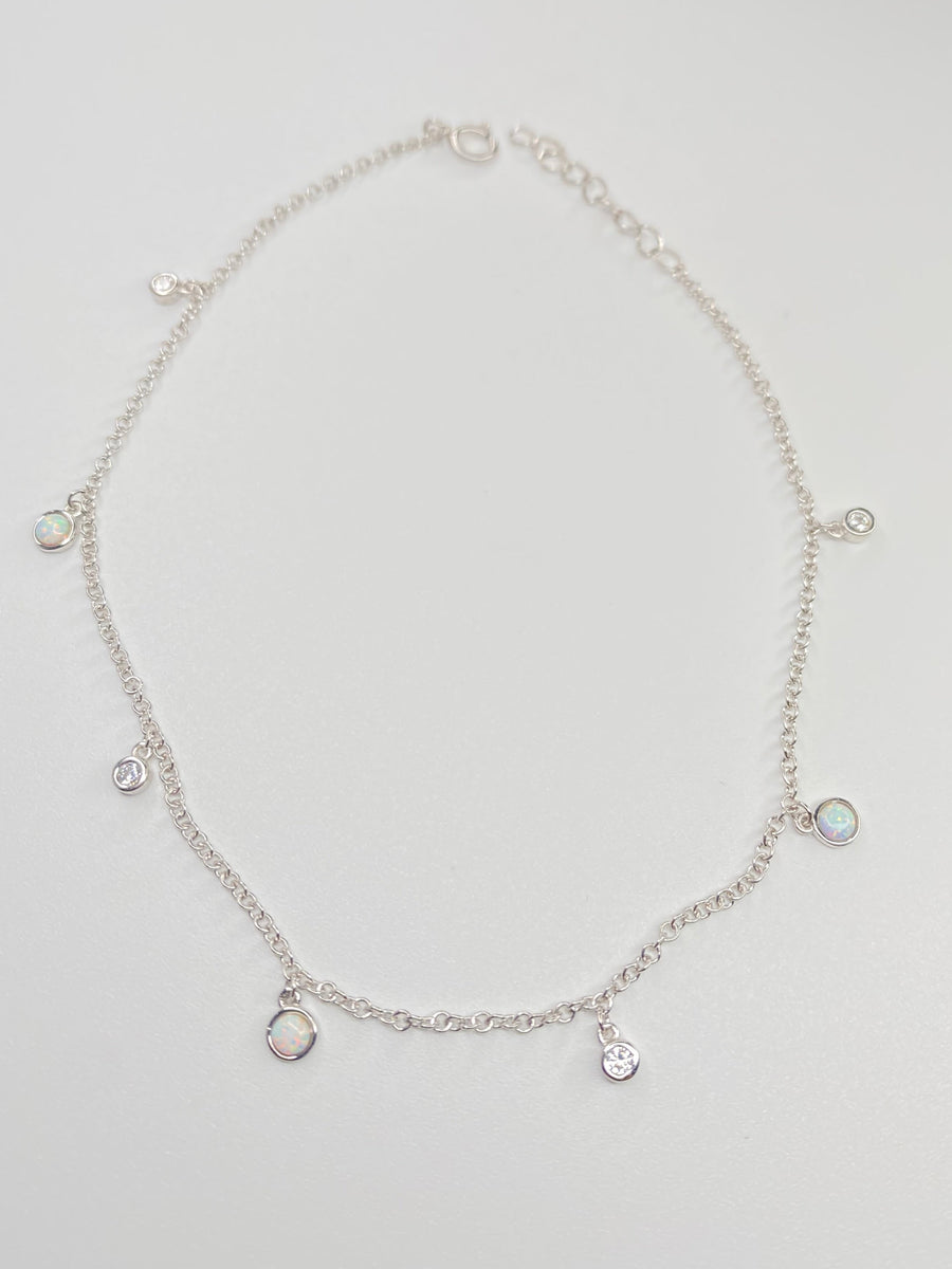 A delicate silver chain anklet with small dangling charms, including a round bezel-set iridescent stone and several clear stones, displayed on a light gray background.