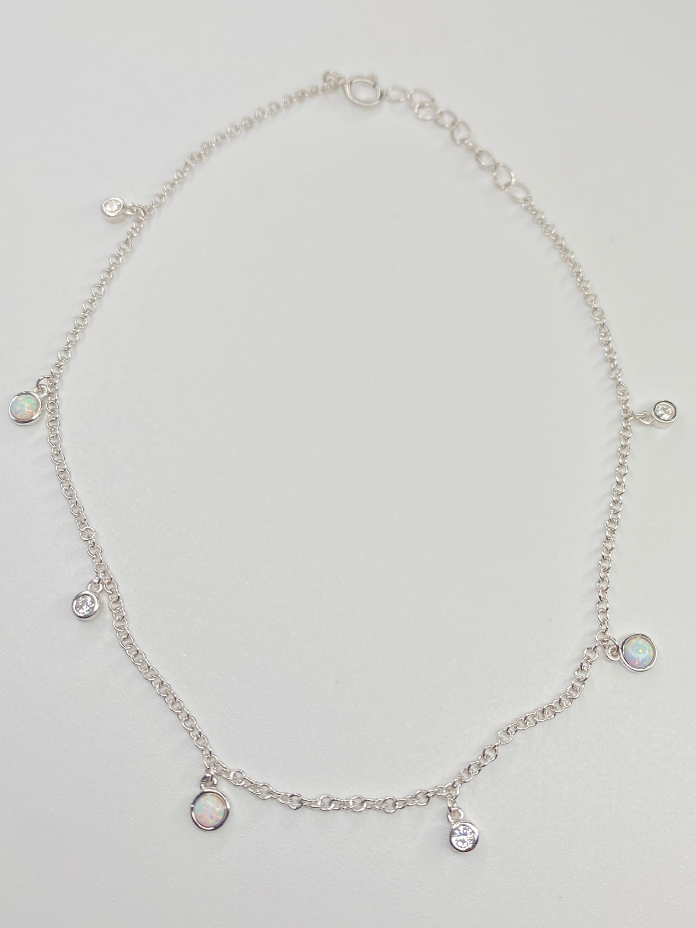 A delicate silver chain anklet with small dangling charms, displayed on a light gray background.