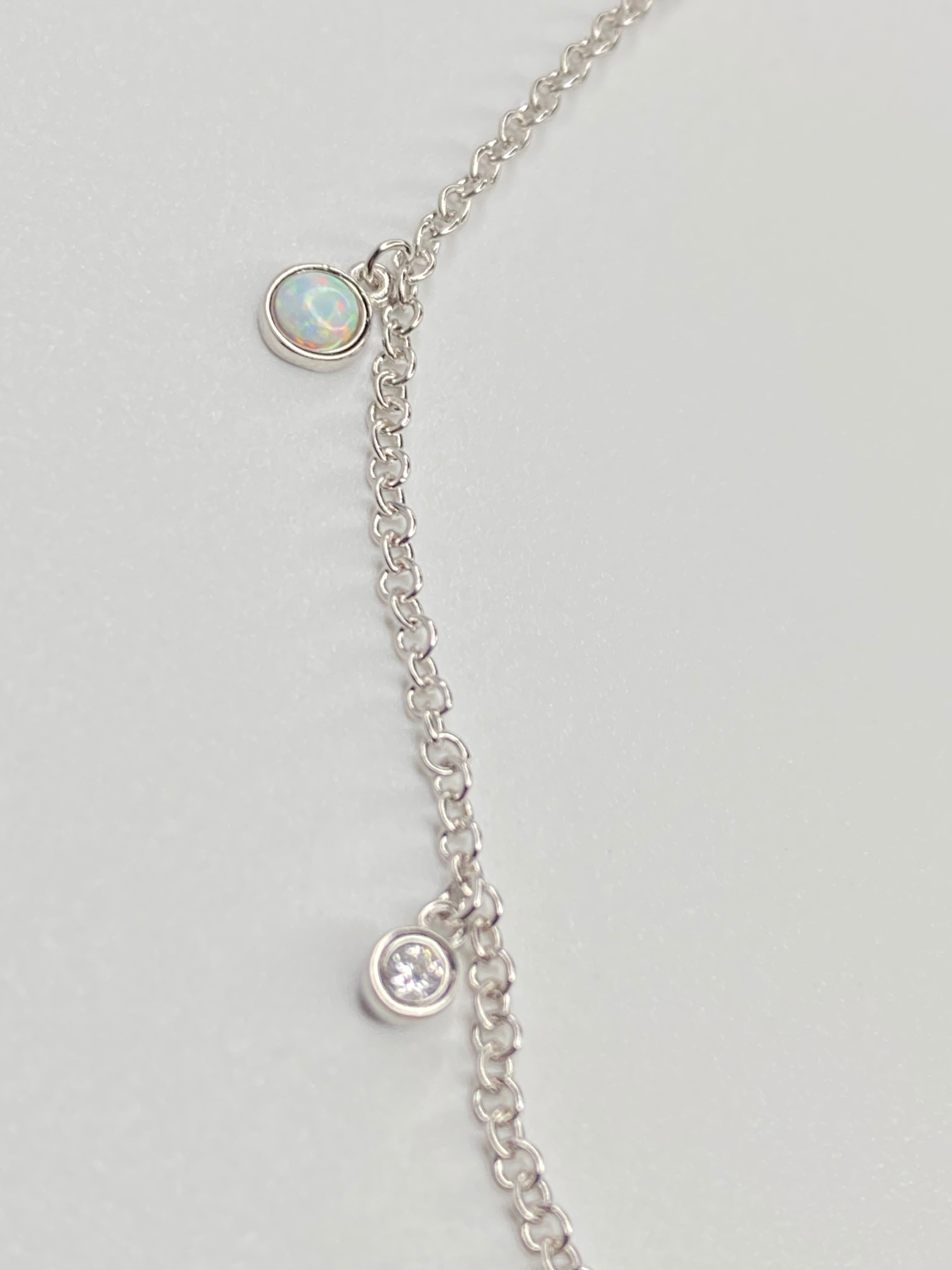 A delicate silver chain necklace featuring two small oval charms, one with a white opal  like stone and one with a clear cz displayed on a light gray background.
