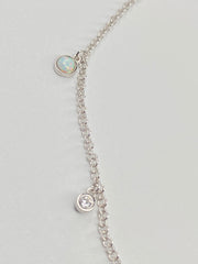 A delicate silver chain necklace featuring two small oval charms, one with a white opal  like stone and one with a clear cz displayed on a light gray background.