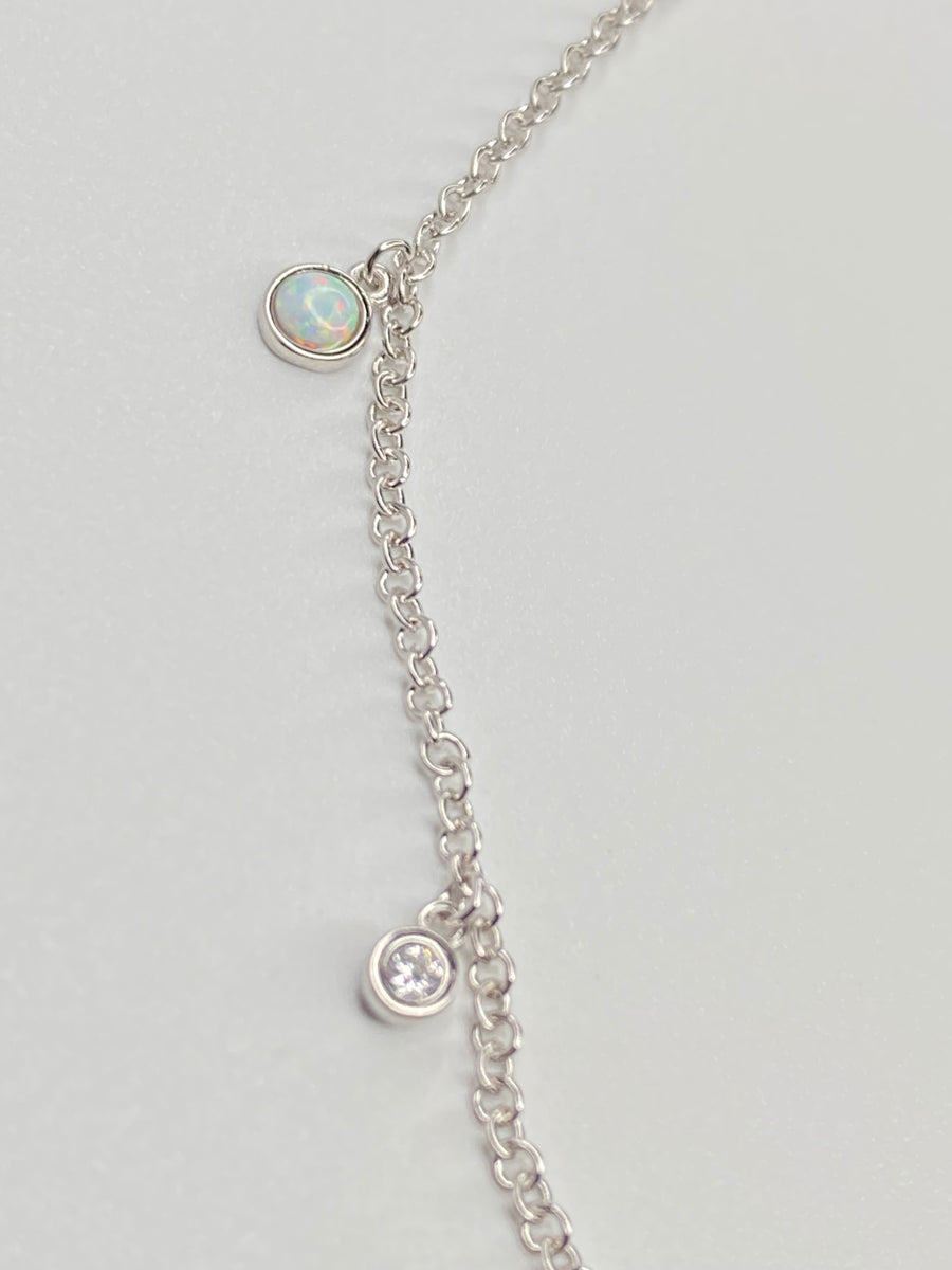 A delicate silver chain necklace featuring two small oval charms, one with a white opal  like stone and one with a clear cz displayed on a light gray background.