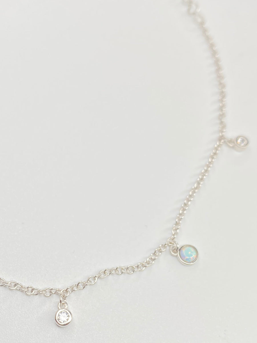 A delicate silver chain anklet with small dangling charms, including a opal like stones and clear cz round stones, displayed on a clean white background.