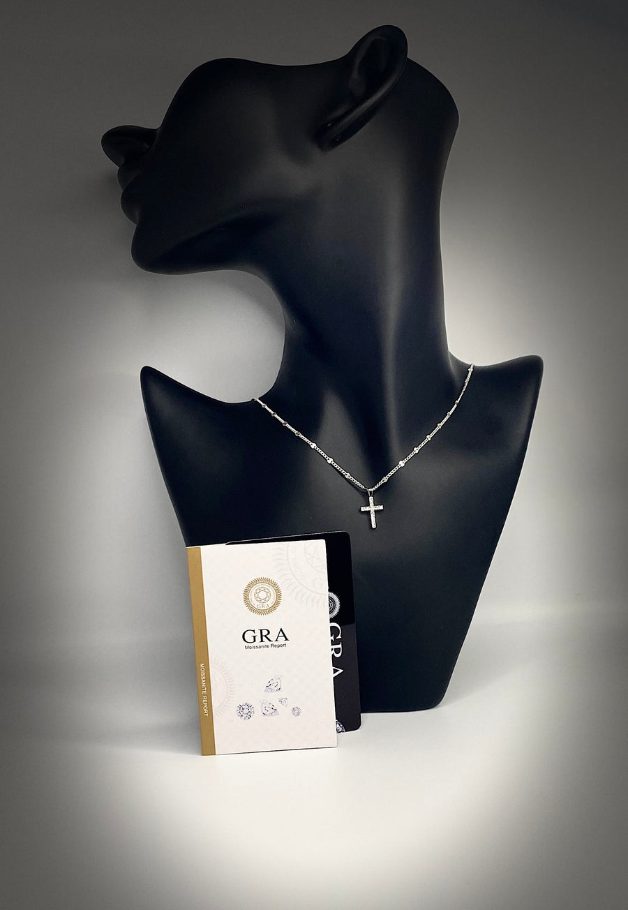 A silver cross pendant necklace displayed on a black mannequin with a moissanite certificate  in front. The bust is set against a dark gray background with dramatic spotlighting.